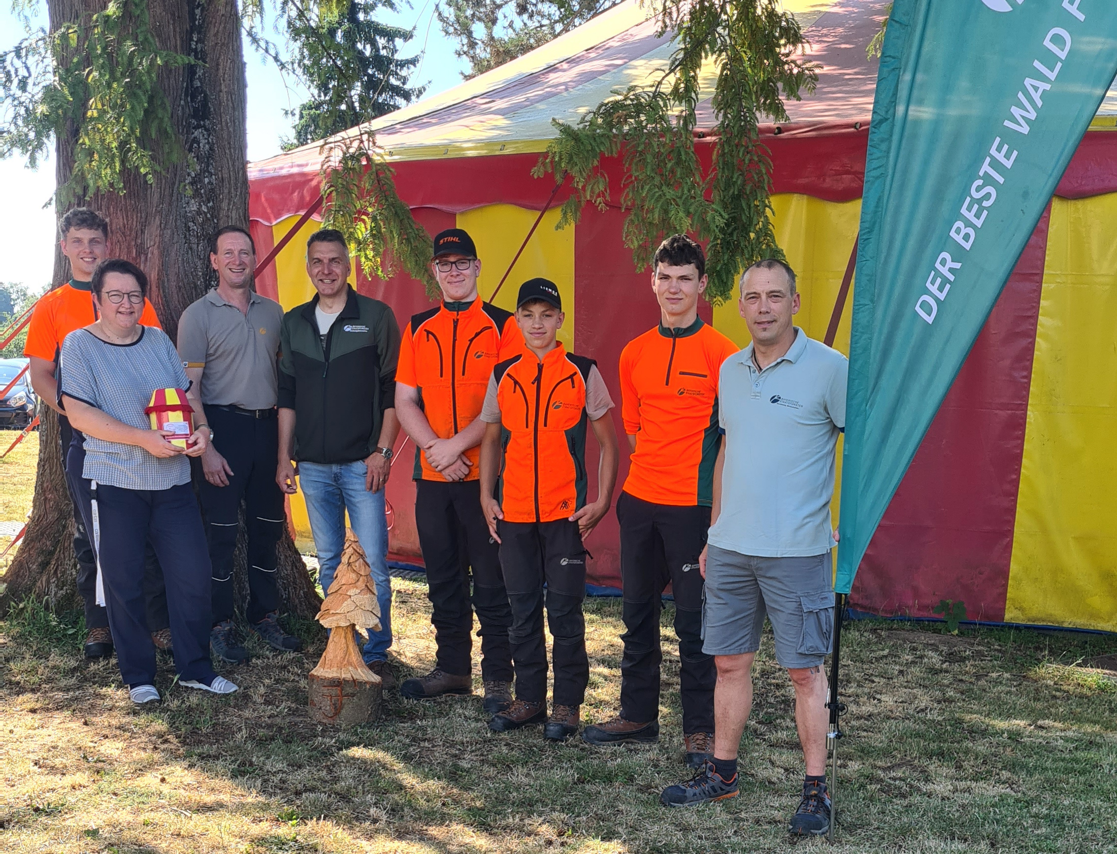 Azubis der Forstbetriebs Arnstein stehen vor einem Zirkuszelt und übergeben die Spende an die Caritas