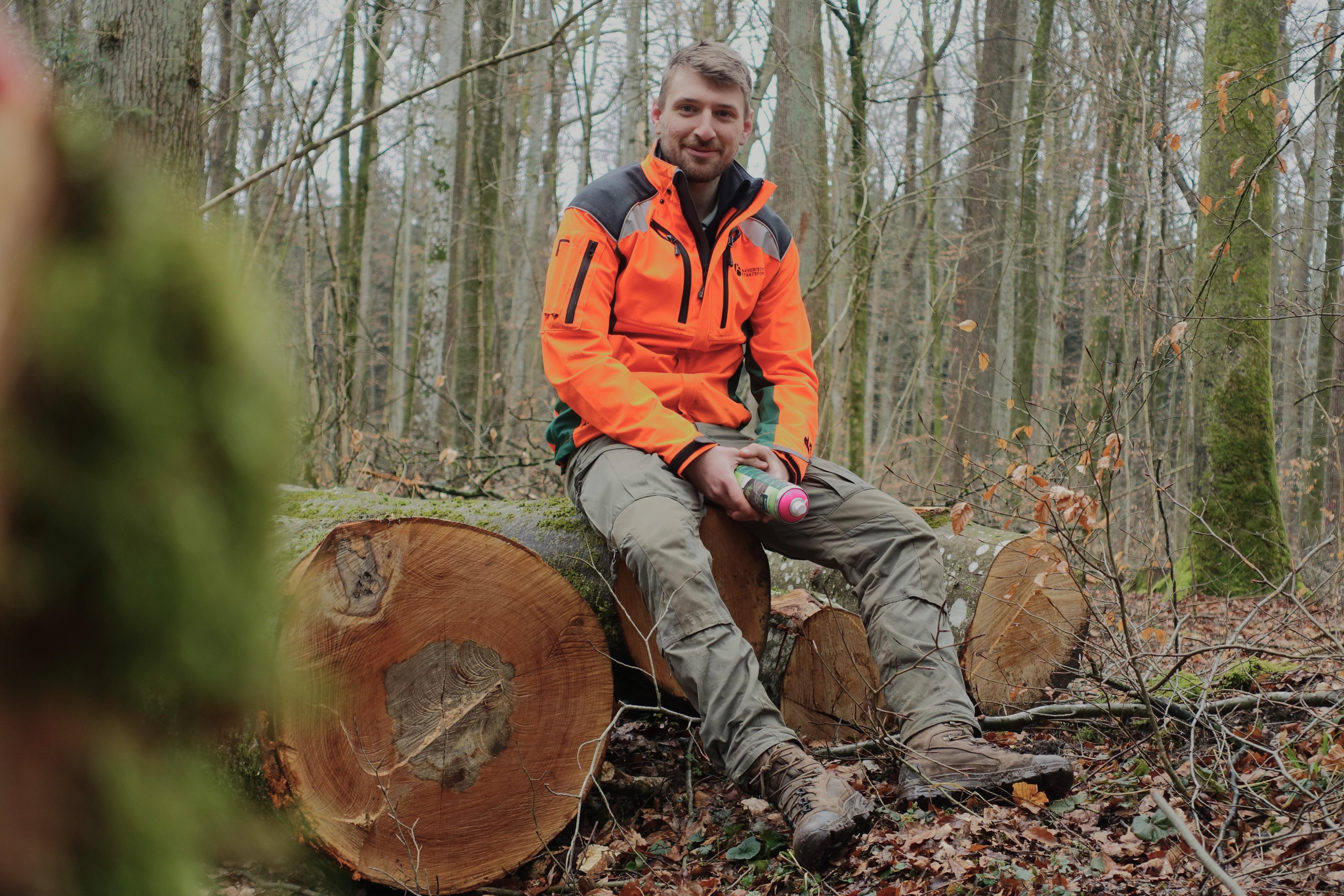 Korbinian Staufer ist neuer Förster im Revier Giesinger Waldhaus in München. Er sitzt auf einem Baumstamm mit einer Spraydose in der Hand